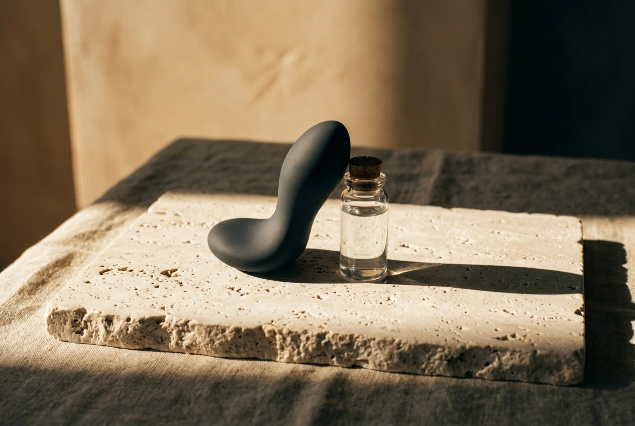 A curved matte black massager resting on a travertine slab beside an amber bottle